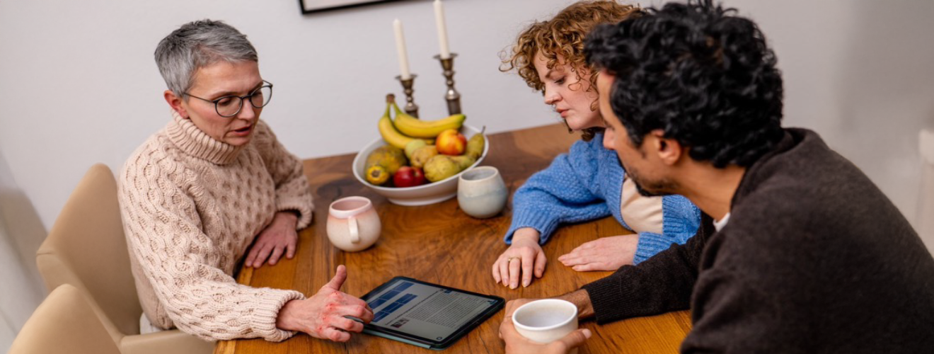 Zwei Frauen und ein Mann sitzen um einen Tisch. Sie besprechen, was sie auf dem Display eines Tablet-PCs lesen.