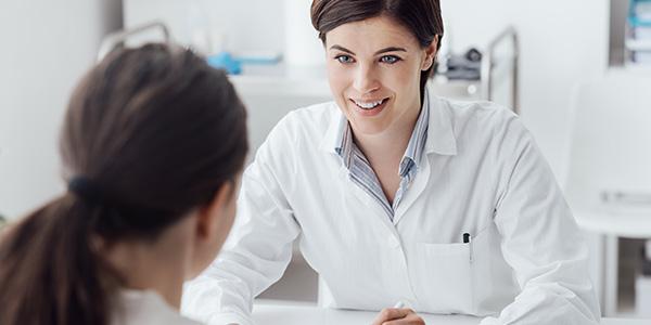 Hier geht es zu den nützlichen Informationen und Materialien Ärztin frontal am Schreibtisch sitzend im Gespräch mit Patientin, von hinten zu sehen.
