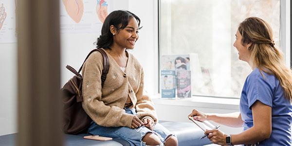 Hier geht es zum Beitrag Ärztin rechts im Bild erklärt weiblicher, junger Patientin, die auf einer Behandlungsliege sitzt, etwas.